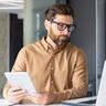 Mann mit Brille hat iPad in der Hand und schaut auf Laptop